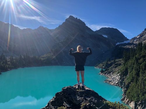 Alexis stands on a rocky peak in the mountains, flexing her arms and smiling, with a beautiful blue lake visible in the background.