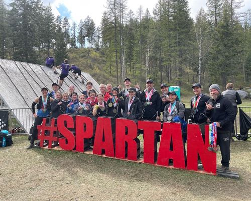 A CoreFit crew of about 20 people hold their finishing medals while standing behind a large sign that says #SPARTAN.